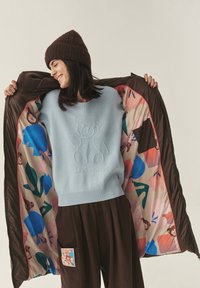 Világoskék kötött pulóver medve mintával, barna steppelt dzseki színes virágmintás béléssel, és barna bő szabású nadrág folttal.