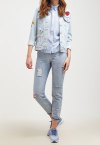 Henkilö, jolla on päällään vaalean denim-takki, jossa on pahvikuvia, vaaleansininen nappipaita, revityt vaaleansiniset farkut sekä siniset urheilukengät.