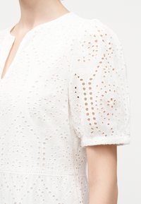 Λευκό μπλούζα από δαντέλα eyelet με φουσκωτά κοντά μανίκια και ελαστικό τελείωμα στο μανίκι, με στρογγυλή λαιμόκοψη και μικρή V-κοπή.