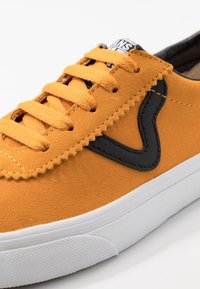 Gros plan sur une basket en daim jaune moutarde avec des lacets assortis, un logo noir sur le côté et une semelle en caoutchouc blanche.