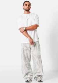 Joven con el cabello trenzado que lleva una camiseta blanca extragrande, pantalones anchos de color gris claro con estampado, zapatillas blancas, joyería plateada y un reloj de pulsera, de pie.