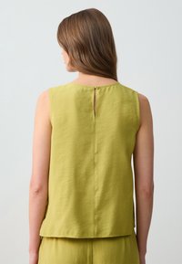 Top senza maniche in tessuto verde chiaro con una finitura texturizzata, caratterizzato da un dettaglio a chiave sul retro e una chiusura a bottone al collo.