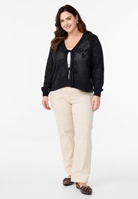 Črn pleteni kardigan z V-izrezom in zavezovanjem, nošen čez belo majico, v kombinaciji s svetlo bež hlačami in balerinkami z živalskim vzorcem.