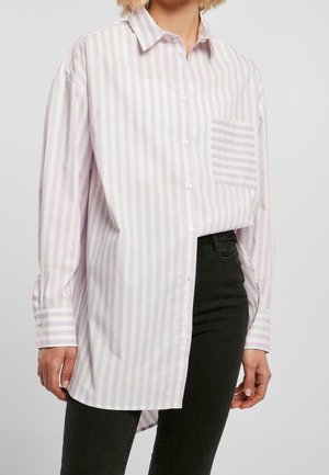 Personne portant une chemise oversize à rayures verticales rose pastel et blanc avec une poche poitrine rayée et un pantalon noir.
