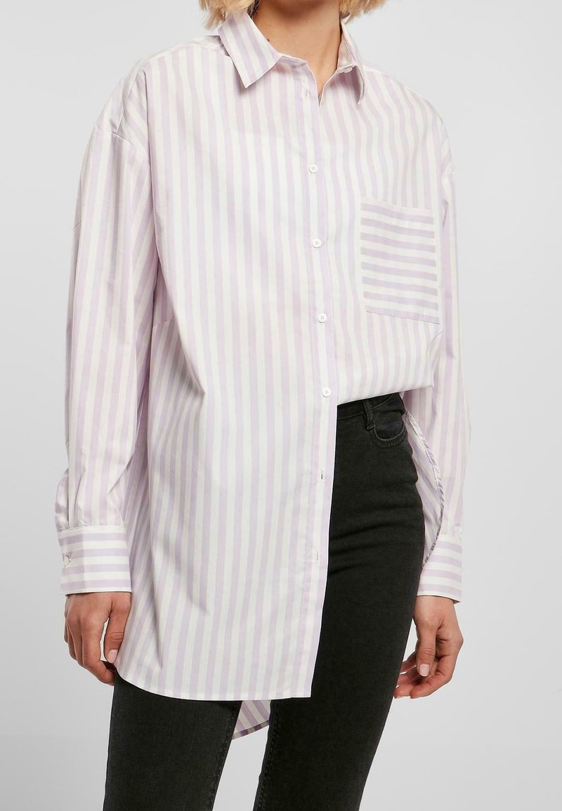 Personne portant une chemise oversize à rayures verticales rose pastel et blanc avec une poche poitrine rayée et un pantalon noir.