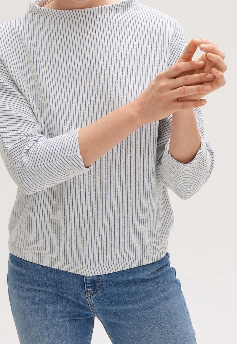 Personne portant une chemise blanche à manches longues avec de fines rayures verticales et un jean bleu, les mains jointes devant le torse.