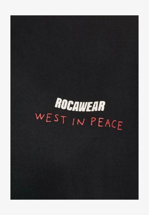 Camiseta de algodón negra con "ROCAWEAR" en una fuente blanca en negrita y "WEST IN PEACE" en letras rojas, posicionado a lo largo del pecho.