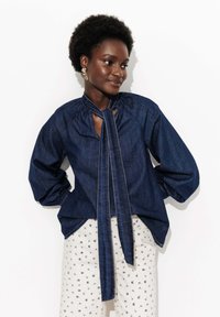 Blouse en denim avec un col à nouer, présentant un décolleté froncé, des manches longues et une coupe décontractée ; associée à un pantalon crème à motifs.