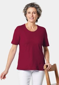 Bordeaux T-shirt met korte mouwen en een ronde hals, gemaakt van zachte stof. Gemaakt in combinatie met witte broek en een houten stoel voor context.