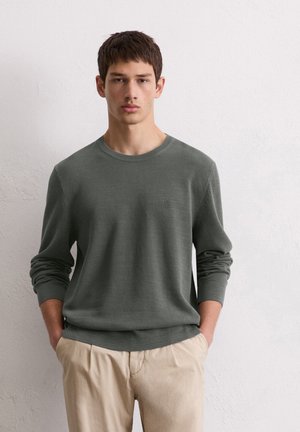 Pull en tricot vert avec un col rond et des poignets côtelés, associé à un pantalon beige clair. Texture subtile sur l'ensemble du tissu.