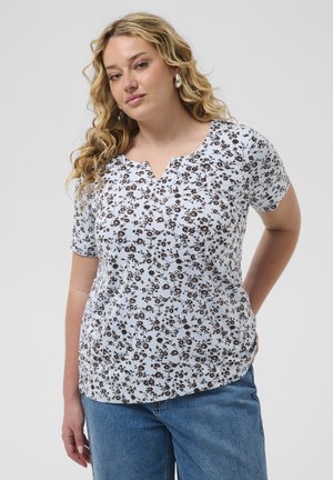 Femme aux longs cheveux blonds bouclés portant un haut blanc à manches courtes à motifs floraux et un jean bleu, debout devant un fond clair uni.
