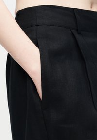 Χέρι που ακουμπά μέσα στην εμπρόσθια τσέπη ενός μαύρου παντελονιού ψηλής μέσης με θηλιές για ζώνη και ραφές προσαρμογής.