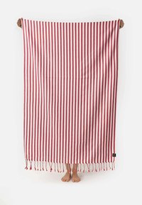 Gestreepte handdoek in rood en wit, met franjes aan de randen. Zachte textuur en lichtgewicht ontwerp, verticaal vastgehouden met blote voeten zichtbaar eronder.