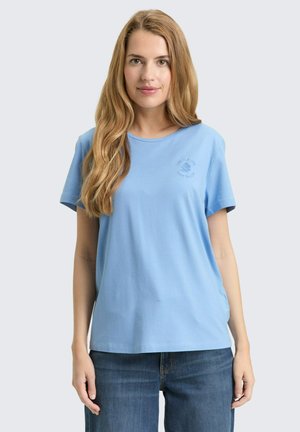 Junge Frau mit langen hellbraunen Haaren, die ein hellblaues kurzärmliges T-Shirt und blaue Jeans trägt, vor einem einfarbigen Hintergrund stehend.