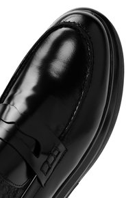 Mocassin en cuir noir brillant avec bout arrondi, coutures visibles et une bride décorative sur la partie supérieure de la chaussure.
