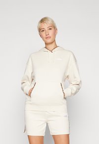 Beige hette med dyreprintdetalj på hetten og lommene, har kengurulomme og logodetalj på brystet. Sammen med shorts.