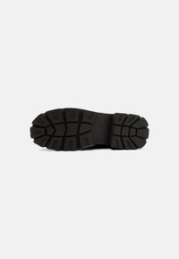 Semelle de chaussure en caoutchouc noir avec des crampons profonds pour l'adhérence, arborant un design structuré et une zone de talon renforcée. Texture lisse dans l'ensemble.