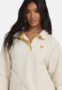 Cremefarbener Fleece-Pullover mit gelben Akzenten, Halbzipp-Kragen und Druckknopfverschlüssen. Ausgestattet mit strukturiertem Material und Seitentaschen.