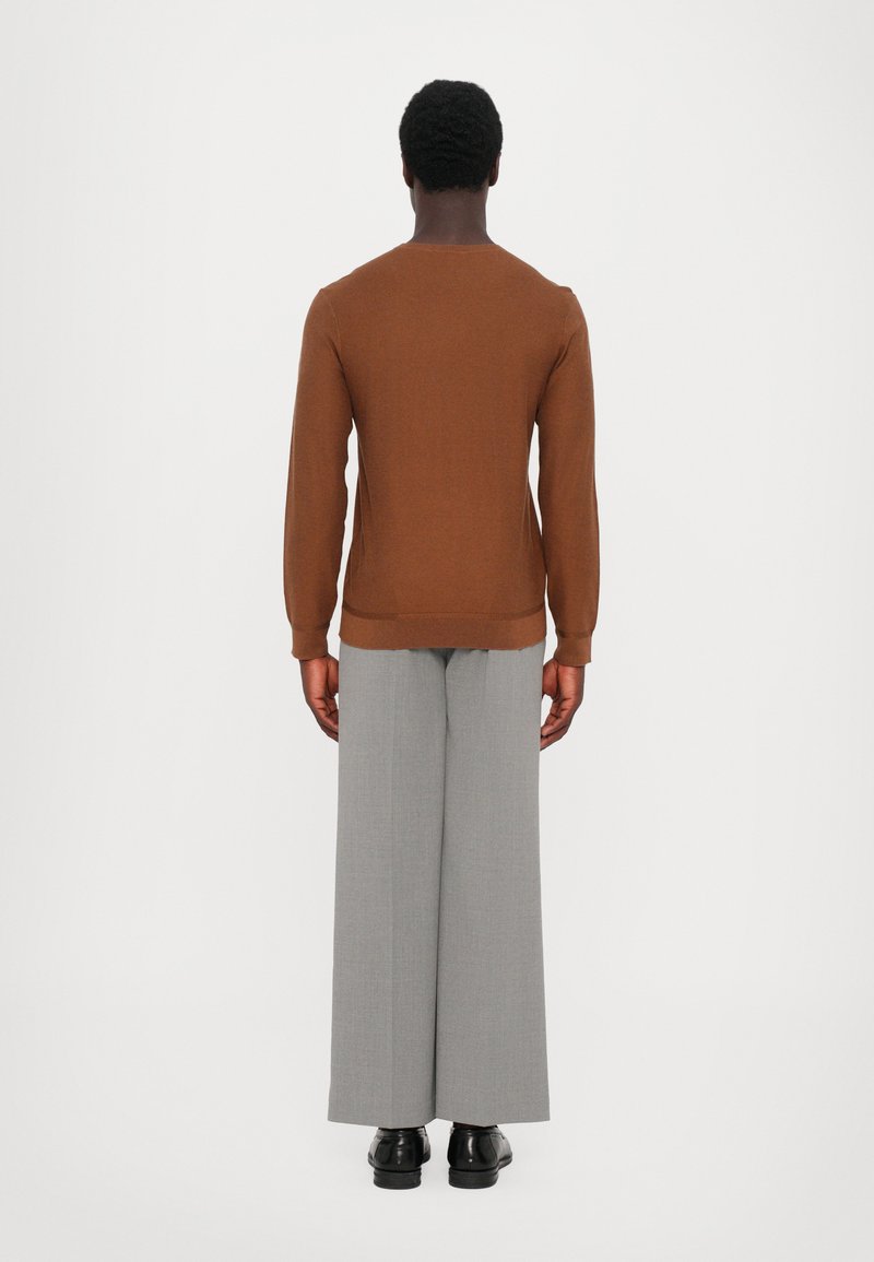 Brun stickad tröja med lång ärm, i kombination med vida grå byxor och svarta skor, sedd bakifrån mot en enfärgad bakgrund.