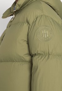 Oliwkowa kurtka puchowa z wysokim kołnierzem i okrągłą naszywką logo Tommy Hilfiger z 1985 roku na górnej części rękawa.