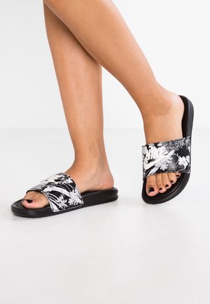 Stopy noszące czarno-białe kwieciste klapki Nike slide z czarnym lakierem do paznokci na palcach, na białym tle podłogi.