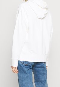 Sudadera blanca con capucha de corte relajado, que presenta una textura suave y puños acanalados. Combinada con jeans de mezclilla azul claro.