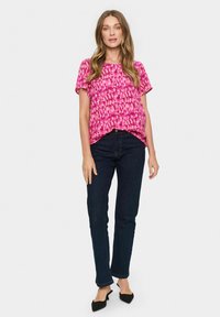 Lyserød kortærmet bluse med et tekstureret mønster, parret med mørke denim jeans. Modellen har spidse sorte flade sko på. En enkel, afslappet outfit.