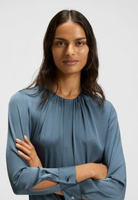 Blouse met lange mouwen in zachtblauwe satijn, met geplooide halslijn en uitgesneden manchetten met ronde knopen. Gladde textuur, aansluitende silhouet.