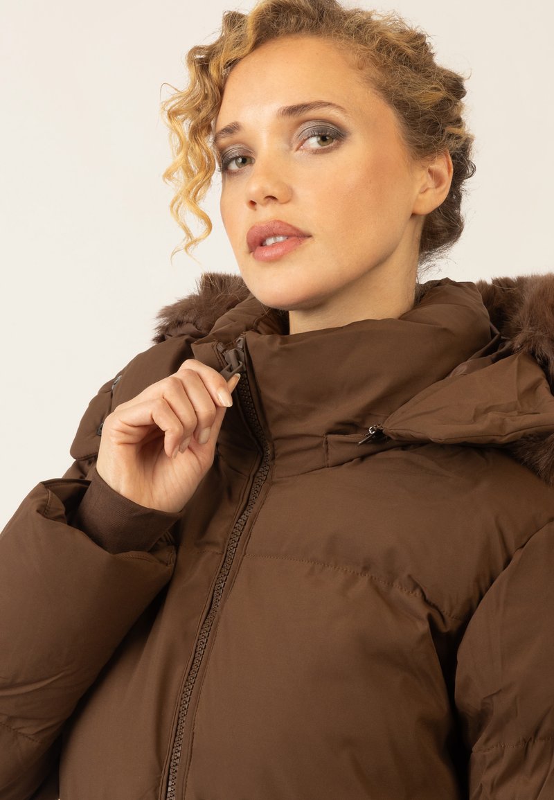 Braune Pufferjacke mit hohem Kragen und Fellbesatz. Verfügt über einen Reißverschluss vorne und eine glatte, matte Textur. Das Modell posiert mit einer Hand auf dem Reißverschluss.