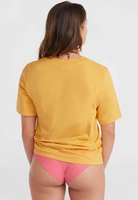 Gul bomulds T-shirt med korte ærmer, glat tekstur, afslappet pasform, båret over lyserøde bikini undertøjsdele, set fra bagsiden.