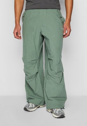 Hellgrüne Outdoor-Hosen mit lockerer Passform, ausgestattet mit einem Kordelzugbund, Taschen und ripstop Textur. Kombiniert mit grauen Wanderschuhen.