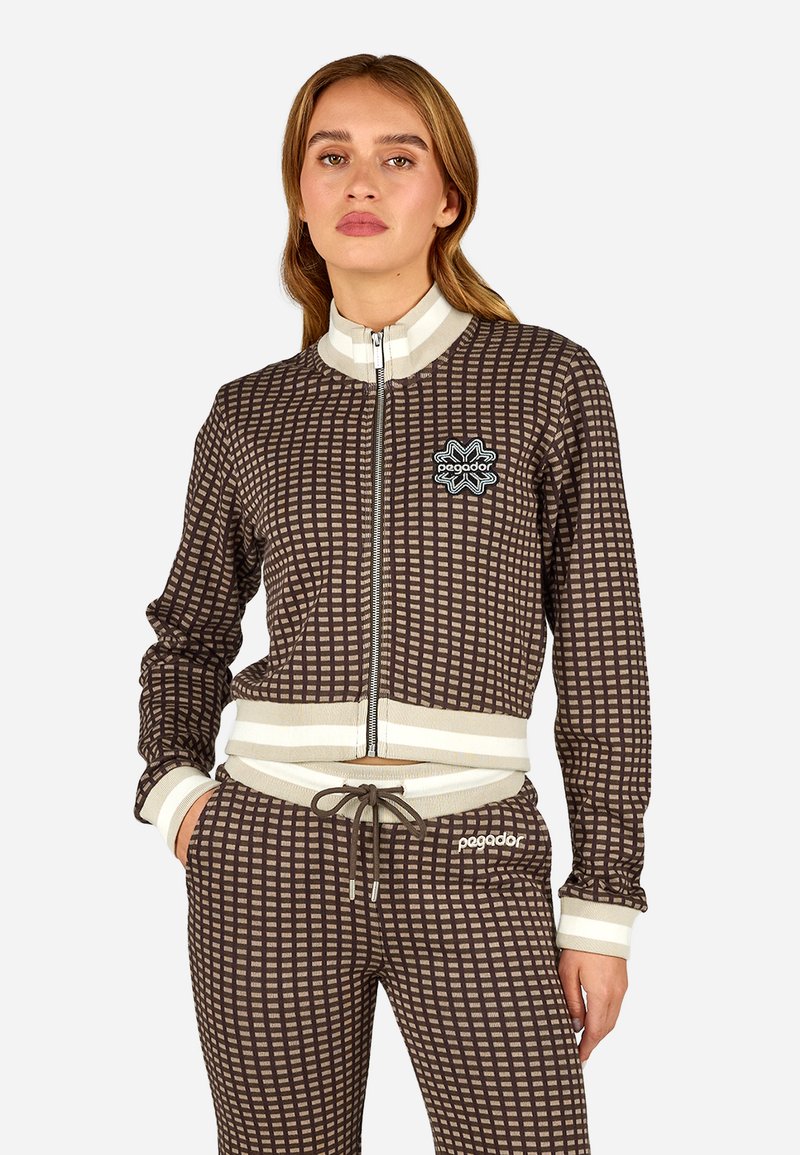 Hnedá károvaná zipsová bunda s rebrovanými bielymi manžetami a golierom, kombinovaná s rovnakými nohavicami s šnúrkou a logom.