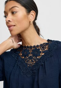 Navy top with intricate floral lace neckline, textured fabric, and loose fit. Gold hoop earring and thin bracelet accentuate the look.