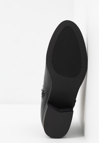Semelle de chaussure noire avec motif de grip texturé au talon et à la zone de la plante du pied, inclinée contre un mur d'angle blanc et un sol.
