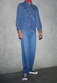 Chemise en denim avec deux poches poitrine, jean bleu et baskets noires avec des accents blancs. La tenue est agrémentée d'un bandana rouge autour du cou.