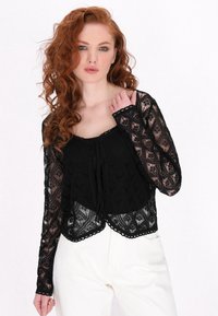 Top din dantelă neagră cu mâneci lungi, având un model geometric, margini ondulate și un sistem de prindere cu fundă în față. Purtați-l peste un strat de bază solid negru.