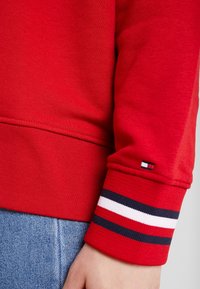 Felpa rossa con polsini a costine, caratterizzata da strisce nere, bianche e rosse. Realizzata in cotone testurizzato, mostrata con un tessuto denim sottostante.