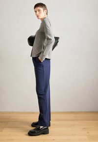 Γκρι εφαρμοστό σακάκι με μια μικρή τσέπη, συνδυασμένο με ναυτικές μπλε παντελόνες και χοντρές μαύρες loafer, σε φωτεινό φόντο.