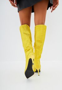 Botine galbene până la genunchi cu un finisaj texturat, vârf ascuțit și toc stiletto. Detaliu cu fermoar lateral pentru o îmbrăcare ușoară.