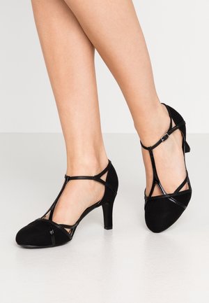 Jambes portant des chaussures à talons hauts en daim noir à bride en T avec une petite boucle sur un fond clair.