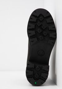 Semelle de botte en caoutchouc noir avec un motif de bande de roulement profond, des rainures horizontales et diagonales, et un accent hexagonal vert près du talon.