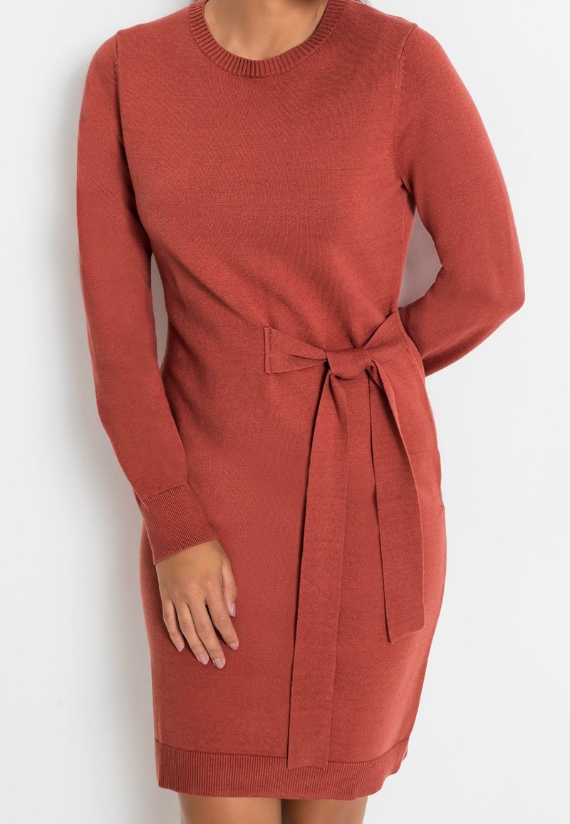 Abito lavorato a maglia color ruggine con maniche lunghe, scollatura rotonda e una cinta in vita. Presenta polsini e orlo a costine per una maggiore texture.