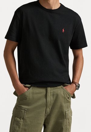 Mann trägt ein schwarzes T-Shirt mit kurzen Ärmeln und rotem Logo, olivgrüne Cargo-Hosen, Hände in den Taschen und eine braune Lederschnalle am Handgelenk.