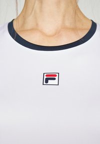 Bílá sportovní košile s námořnickým límcem, která má malé červené a námořnické logo. Materiál má hladkou texturu a splývavý střih.