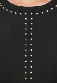 Černý látkový top svisle zdobený řadami zlatých cvoků vpředu. Kulatý výstřih s hladkou texturou a přiléhavým střihem.