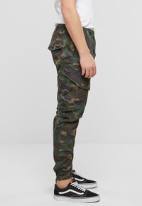 Pantalones cargo de camuflaje en verde y marrón con cintura elástica, bolsillos laterales y puños elásticos. Combinados con zapatillas negras.