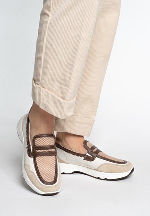 Beige and white slip-on shoes feature mesh and suede materials, brown accents, a rounded toe, and a textured sole. Worn with cuffed beige pants.