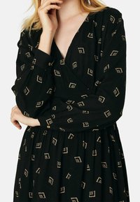 Robe noire à manches longues, décolleté en V et motif carré doré. Présente un tissu texturé et une taille cintrée, accentuée par des détails de boutons.