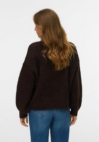 Frau mit welligem, hellbraunem Haar, die einen dunkelbraunen strukturierten Pullover und Bluejeans trägt, steht mit dem Rücken zu einem schlichten Hintergrund.