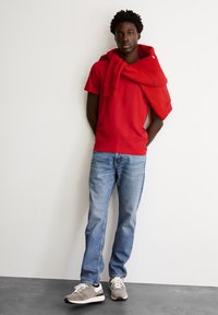 Rode T-shirt met korte mouwen, met een rode hoodie over de schouders, gecombineerd met lichtblauwe jeans en grijze sneakers met witte accenten.